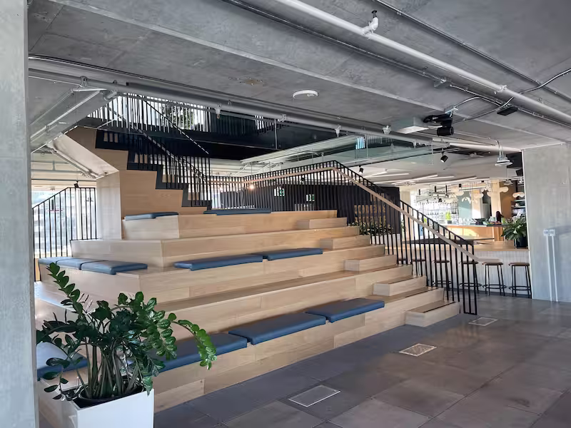 A modern interior space featuring tiered wooden stadium-style seating with grey cushions arranged in ascending steps. The seating area is flanked by black metal railings and has an industrial-style exposed ceiling with visible ductwork and lighting. A large potted plant with broad green leaves sits in the foreground. The space has a minimalist design with concrete flooring and transitions into what appears to be a bar or counter area visible in the background. The overall aesthetic combines warm wood tones with industrial elements and natural accents.